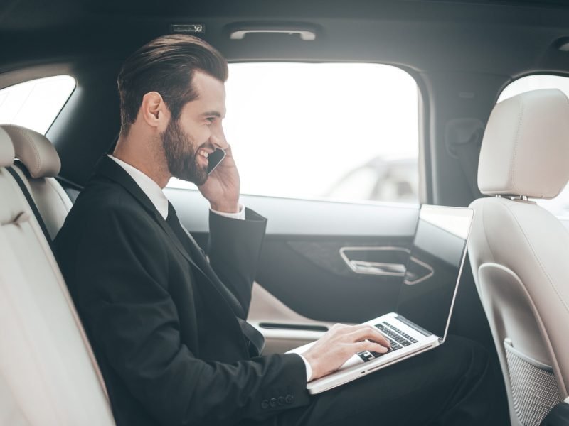 Time to make decision. Confident young businessman working on his laptop and talking on the phone while sitting in the car